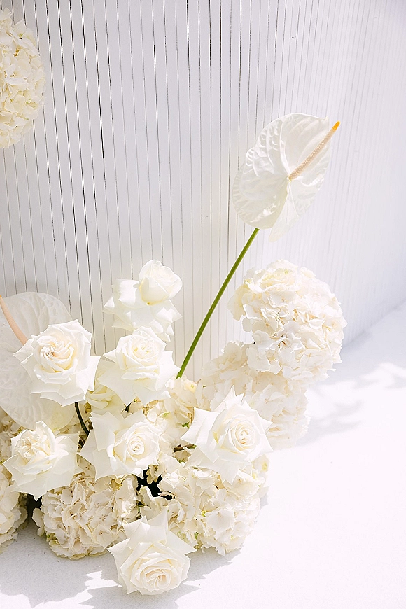 Wedding floral arrangement with white wedding flowers—roses, hydrangea, and anthurium with greenery on a white paneled wall floor in natural light