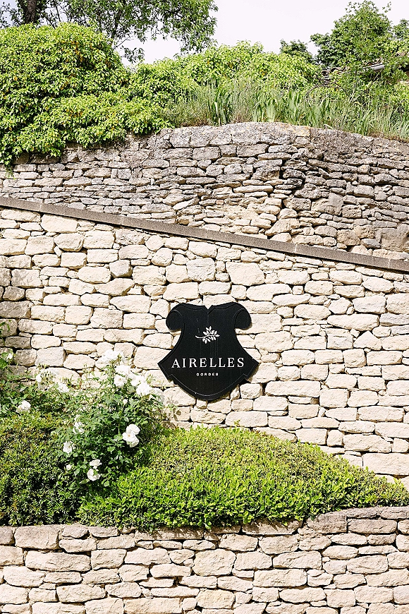 Wedding venue sign with a black plaque and white flowers set against a rustic stone wall with hedges, garden greenery, and sky