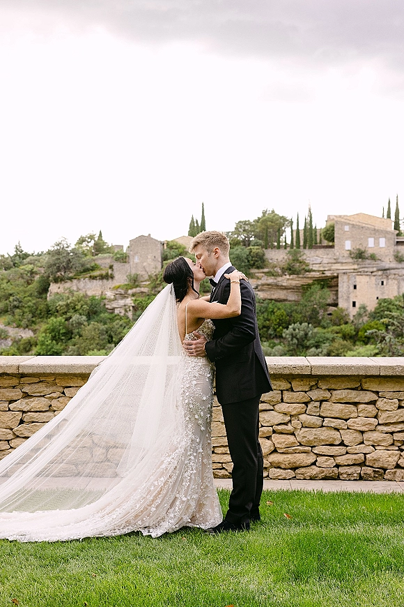 Wedding kiss portrait of bride and groom kissing, veil blowing over lace dress beside stone wall with cypress trees and hillside village beyond