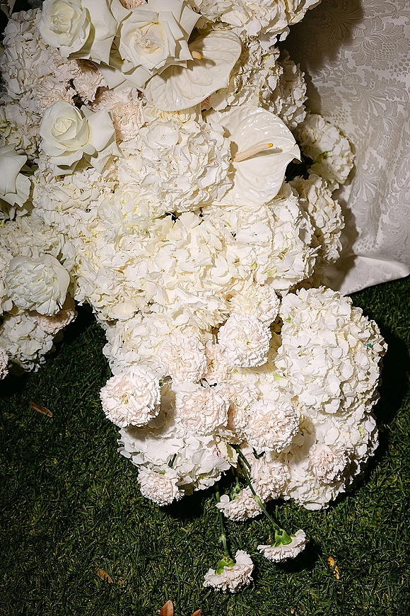 Wedding bouquet of white roses and hydrangeas with anthurium accent, tied with ribbon, resting on grass by patterned fabric backdrop