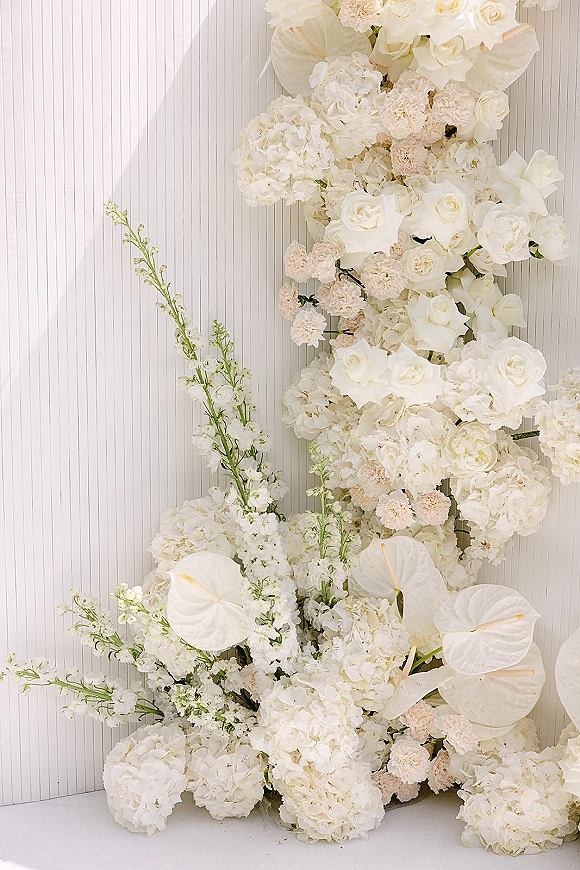 Wedding floral installation with white floral wedding backdrop of roses, hydrangeas, blush carnations and anthuriums against a white paneled wall