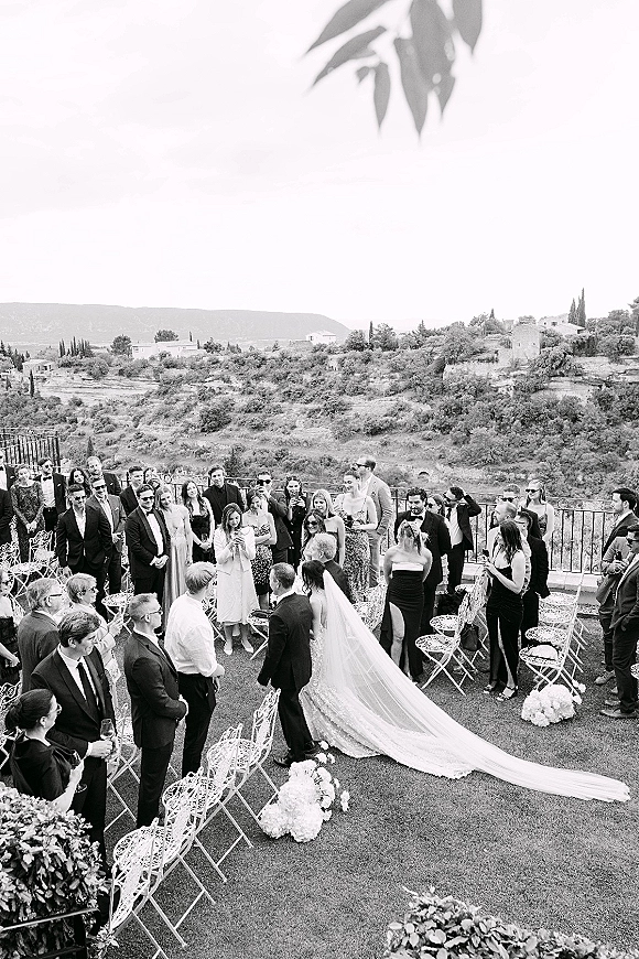 Wedding ceremony moment outdoors as the bride walks down the aisle with a long veil and train, guests in suits watching on a terrace with hillside view