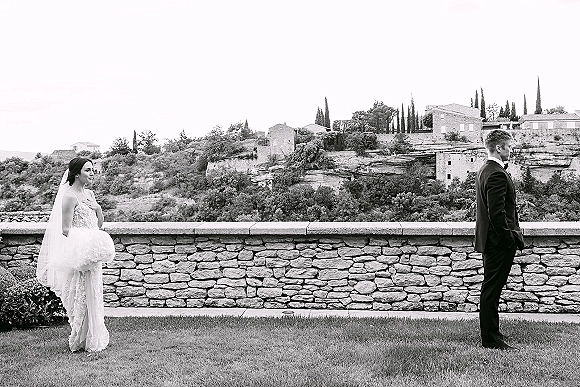 First look moment as bride in wedding dress and veil approaches groom in tuxedo with bow tie by a stone wall on a hillside lawn