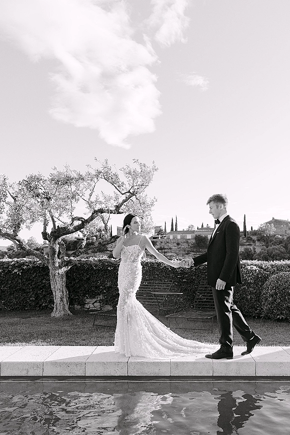 Couple portrait, black and white wedding portrait of bride in strapless lace gown with long train and groom in tux by a poolside lawn