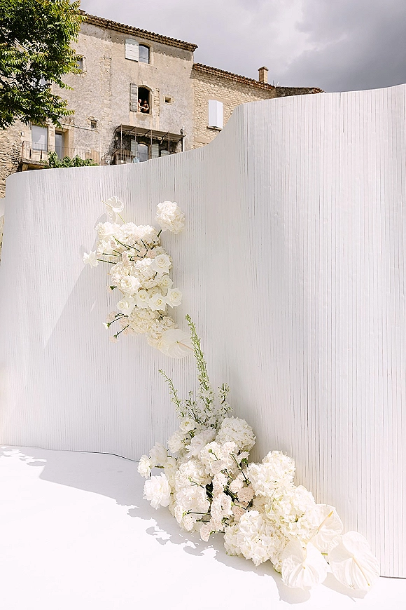 Wedding ceremony backdrop with white ceremony backdrop panels and asymmetrical roses, hydrangeas, anthurium, and greenery by a stone building outdoors