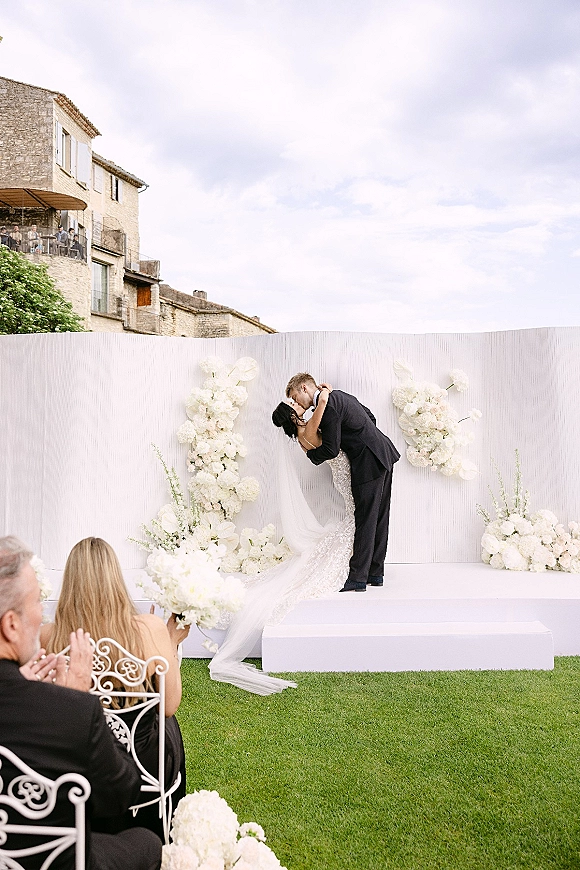 Ceremony kiss as bride in lace dress and veil kisses groom in suit beneath a white floral arch on a lawn by a stone villa