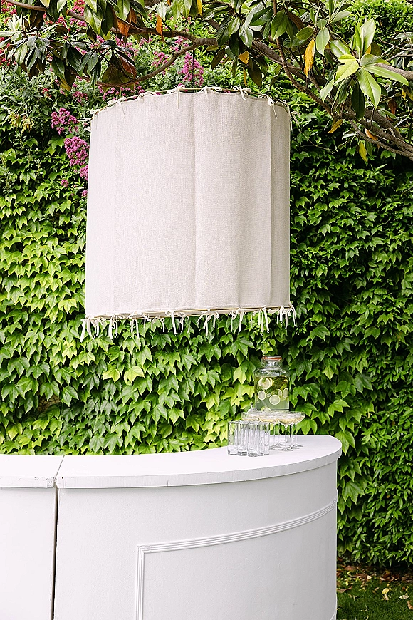 Drink station setup with wedding drink station styling on a white curved bar, citrus dispenser jar and coupe glasses against an ivy wall backdrop.