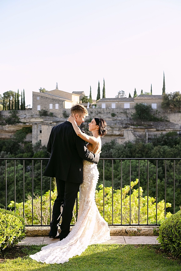 Couple portrait of bride in a strapless lace gown and groom in a tuxedo embracing on a garden terrace with stone villas and cypress trees
