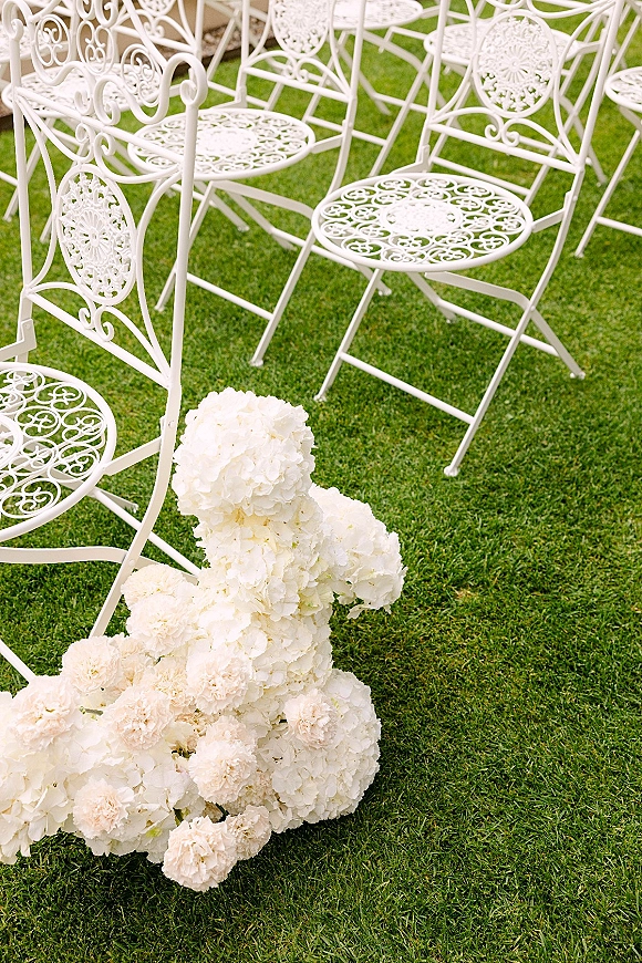 Ceremony seating with outdoor ceremony chairs lined on a green lawn, paired with a ground floral arrangement of white hydrangeas and carnations