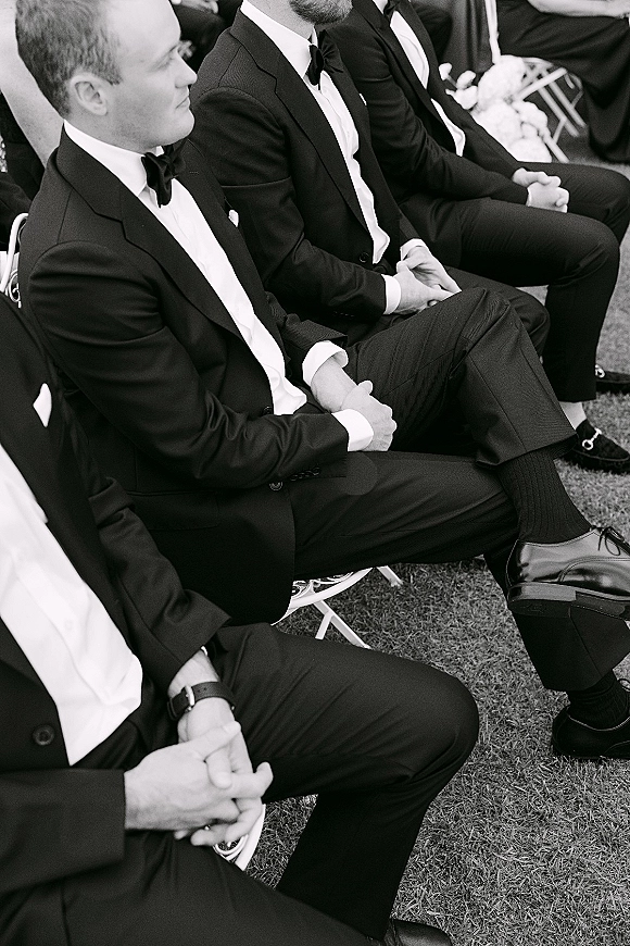 Groomsmen ceremony scene with men in black tuxedos and bow ties seated on white folding chairs on a grass lawn with guests behind