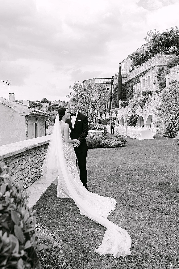 Couple portrait of bride in a lace wedding dress with long veil holding hands with groom in tuxedo on a garden lawn by ivy arches