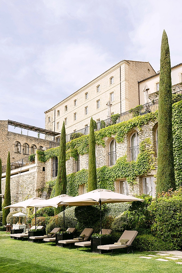 Wedding venue exterior with ivy-draped stone villa, cypress trees, and garden umbrellas over lounge chairs on a manicured lawn terrace