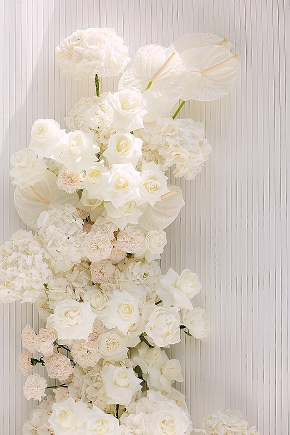 Wedding floral arrangement of white wedding florals with roses, anthurium, hydrangea and carnations against a white ribbed wall backdrop