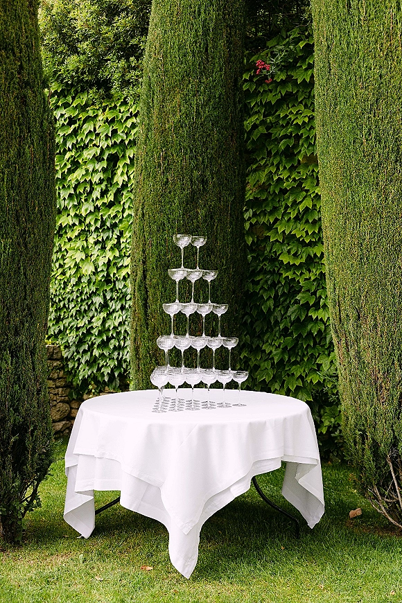 Champagne tower setup with a champagne coupe tower on a round white tablecloth table, set on a garden lawn by an ivy stone wall