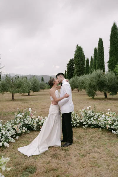 Wedding kiss portrait of bride and groom kissing outdoors, strapless gown and bow tie beside ground florals on an olive grove lawn