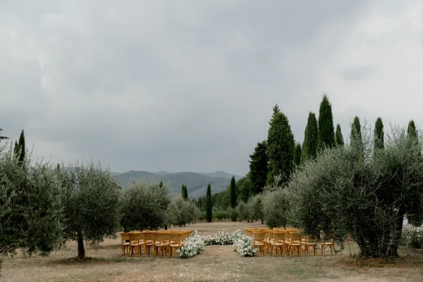 Outdoor ceremony setup with garden wedding ceremony aisle flowers, wood chairs and white florals in a grassy field with olive trees and mountains