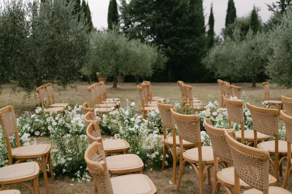 Ceremony aisle decor with outdoor ceremony aisle flowers, low white and blush florals with greenery beside wood and woven chairs in an olive grove lawn