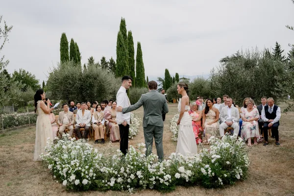 Wedding ceremony outdoors with bride and groom at altar, officiant and guests seated beside white florals and greenery on a lawn