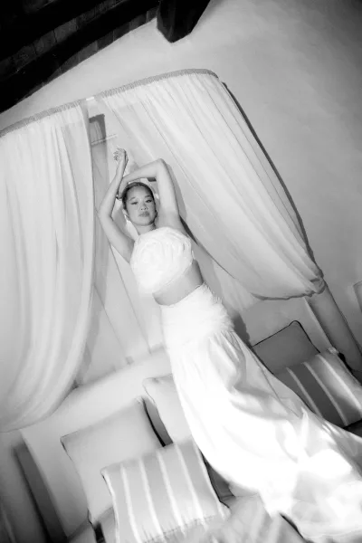 Bridal portrait in a strapless wedding dress, bride with arms raised on a sofa by sheer curtains in an indoor room with window