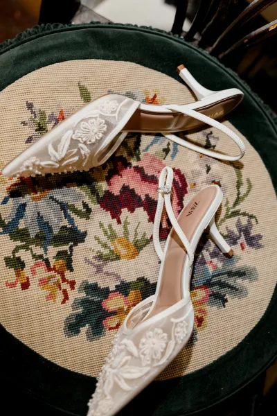 Wedding shoes, white slingback bridal heels with floral embroidery and beaded details, perched on a dark green velvet needlepoint chair seat