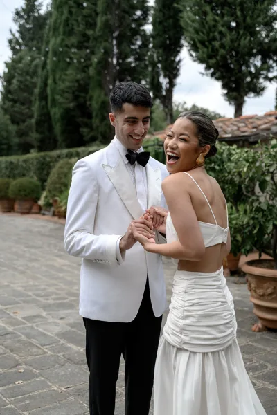 Couple portrait of bride and groom holding hands, laughing in an outdoor courtyard, groom in white tuxedo jacket and black bow tie