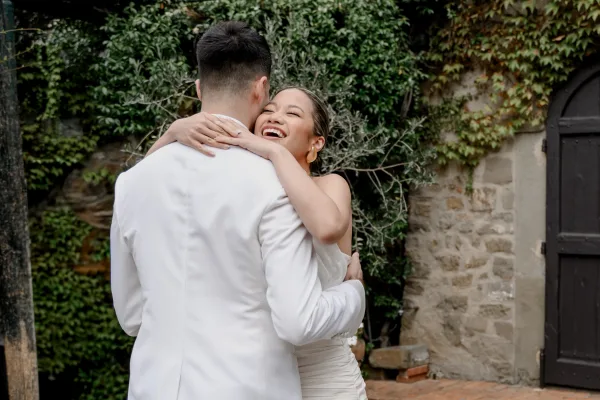 Wedding couple portrait of bride and groom hug, her strapless dress and gold hoops visible as they laugh by an ivy-covered stone wall