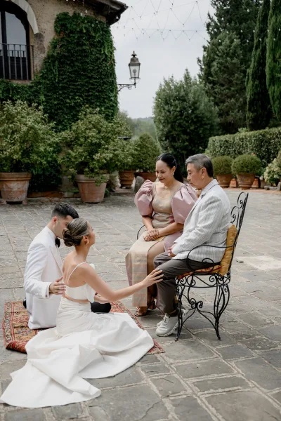 Wedding family portrait of newlyweds with parents in a stone courtyard, bride seated and groom kneeling under string lights by an ivy wall