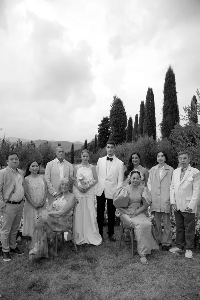 Wedding family portrait with bride holding a bouquet and groom in a white tuxedo jacket, surrounded by relatives on a garden lawn with mountains beyond