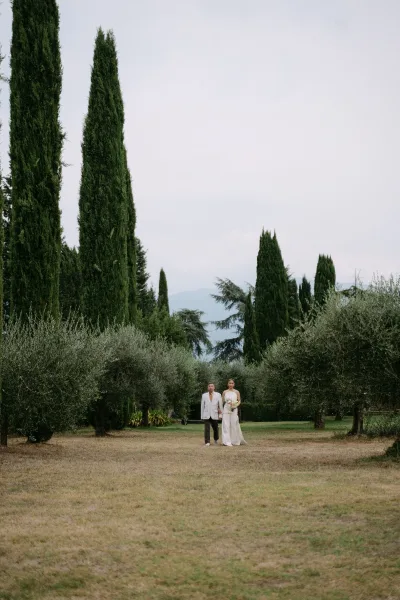 Couple portrait of bride and groom walking hand in hand, bride holding bouquet in a grassy field with cypress trees and mountains beyond