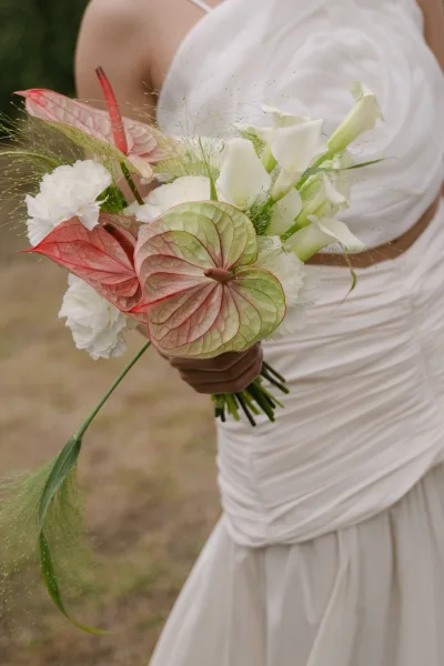 Bridal bouquet with calla lily bridal bouquet blooms and greenery, held against a white wedding dress on an outdoor lawn with blurred foliage