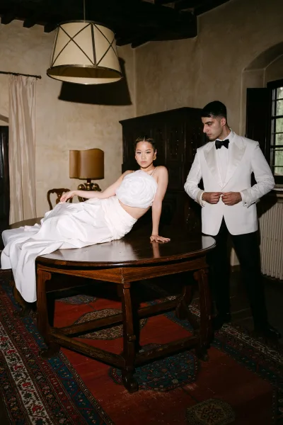 Couple portrait with bride on table pose in a bridal two piece outfit beside groom in white tuxedo jacket in rustic, moody interior