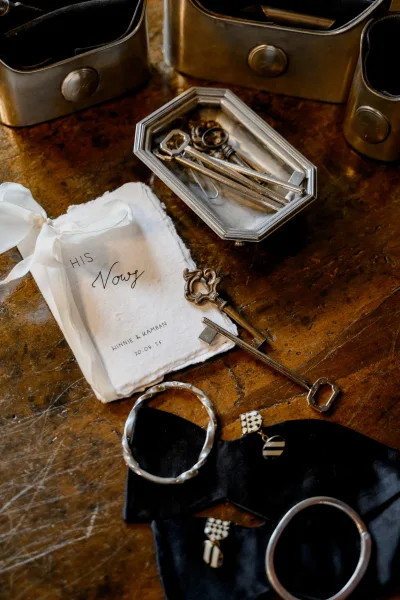 Wedding vow book flatlay with linen vow booklet and ribbon tie beside antique keys and jewelry on a rustic wood tabletop