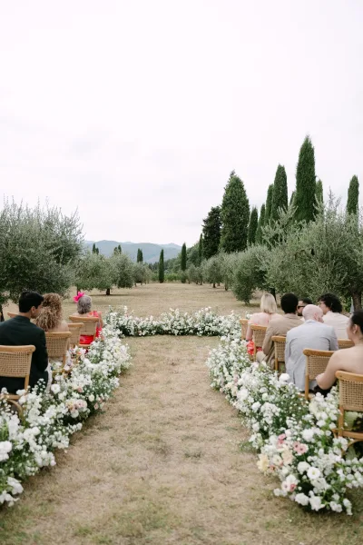 Outdoor ceremony setup with wedding aisle florals lining a grassy aisle between wicker chairs, set in an orchard with mountains beyond