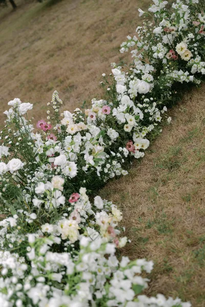 Ceremony aisle flowers in white and blush with greenery accents arranged low along a grass lawn for an outdoor wedding aisle moment