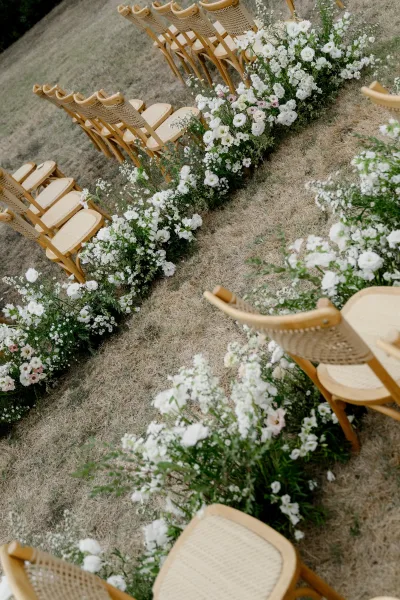 Ceremony aisle decor with outdoor ceremony aisle flowers, white blooms and greenery edging the path beside wood and cane chairs on dry grass lawn