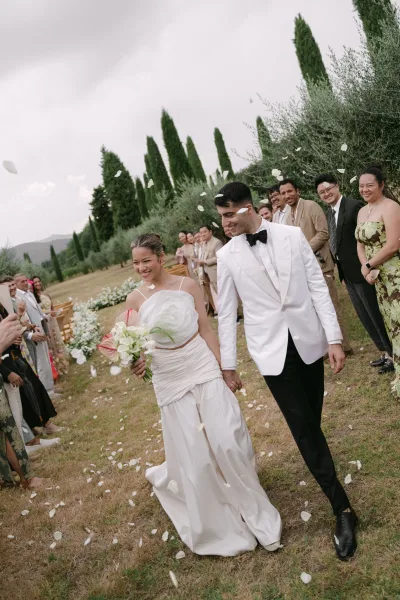 Wedding recessional with bride and groom walking aisle as guests toss rose petals, bride holds bouquet under cloudy sky in garden