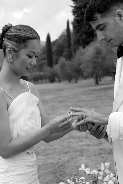Ring exchange as the bride places a wedding ring exchange moment on the groom’s hand, with cloudy garden backdrop and her strapless gown visible