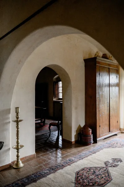 Wedding venue interior with an arched doorway, brass floor candlestick, wooden armoire, vintage chairs, and patterned rug under wood beams