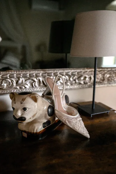 Bridal shoes with lace wedding shoes detailing, ivory pointed-toe slingback heels on a wooden tabletop beside a cat figurine and mirror