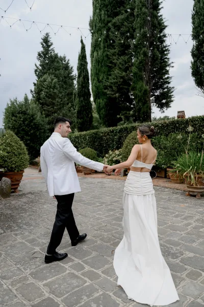 Couple portrait of newlyweds holding hands, walking away in a stone courtyard with string lights, cypress trees, and potted plants under clouds