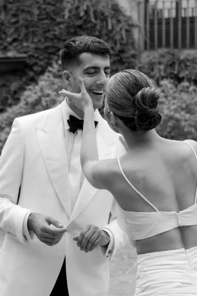 Couple portrait in a black and white wedding portrait, bride touching groom’s face as he smiles, his white tuxedo jacket by garden greenery