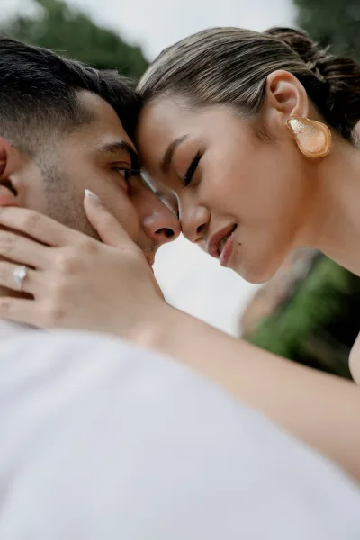 Wedding couple portrait close up of bride and groom forehead-to-forehead, her hand on his face showing an engagement ring against blurred greenery