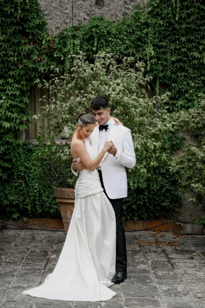 Couple portrait of bride and groom embrace beside an ivy wall, bride in a strapless ruched gown with train and groom in a white tuxedo jacket