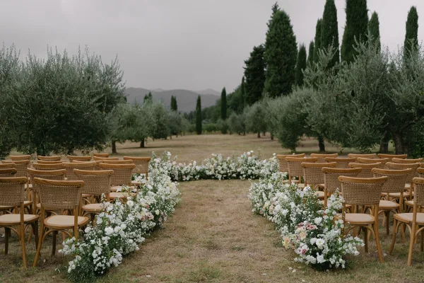 Outdoor ceremony setup with ceremony aisle flowers lining a grass aisle between wood chairs, set in an olive grove under overcast skies