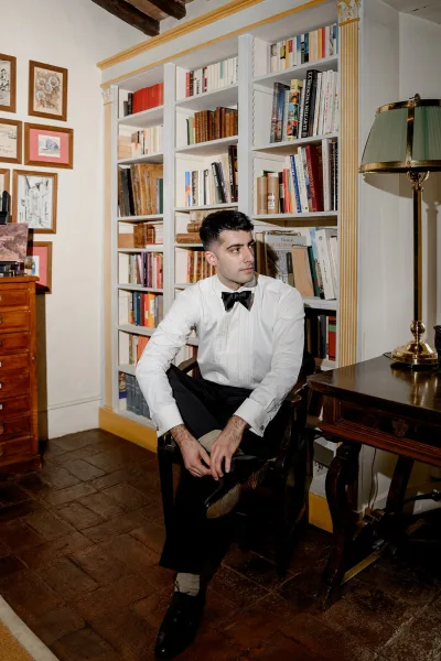 Groom portrait with a black bow tie, seated in a wooden chair, wearing a white tuxedo shirt in a library with bookshelves and art