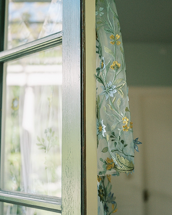 Wedding dress detail with floral embroidered wedding dress lace on sheer tulle, softly lit by natural light beside a window frame