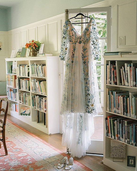 Wedding dress hanging in a library room, embroidered floral long sleeves and tulle skirt by bookshelves, with bridal shoes below
