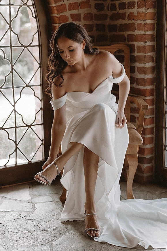 Bride getting ready in an off the shoulder wedding dress, seated on a wooden chair as she slips into rhinestone ankle strap heels by a leaded glass window