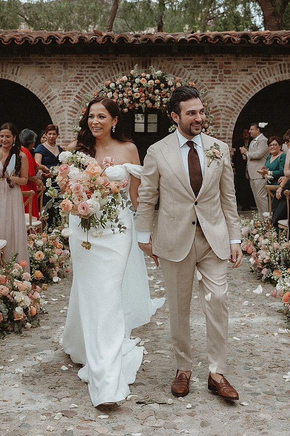 Wedding recessional as bride and groom walk hand in hand down a rose petal aisle, bride holding pastel bouquet in a brick-arch courtyard