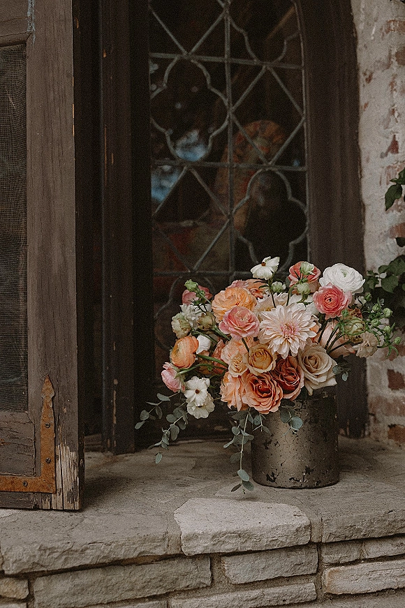 Wedding floral arrangement of pastel roses, ranunculus, and dahlias in a metal bucket on stone steps by a rustic wooden door
